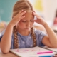 Dolore testa bambini scuola, mal di testa ragazza studio, salute e cura bambini, gestione stress scolastico, supporto medico pediatrico per mal di testa.